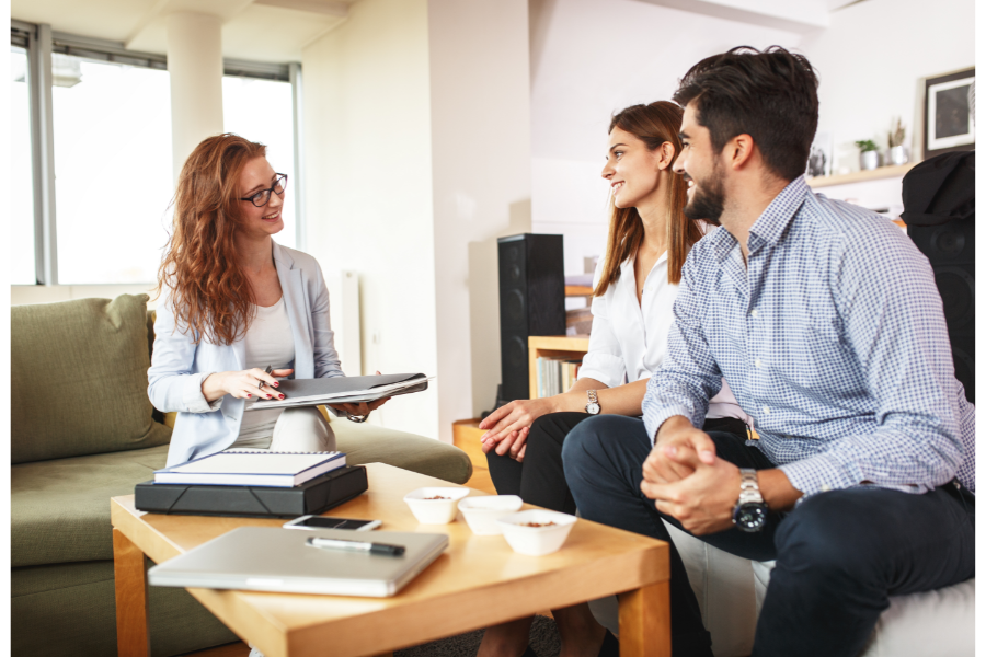 a couple sitting on a couch talking to a real estate agent over paperwork